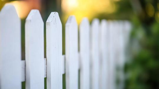 Valkoinen puuaita lähikuvassa, jossa keskimmäinen lauta on selvästi vinossa ja hieman irronnut paikoiltaan. Taustalla näkyy syvyysterotettu puutarhaympäristö.